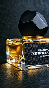 A close-up of a Resonance Eau de Parfum bottle with a faceted black cap and amber-colored liquid inside a clear glass bottle, set against a dark textured background.