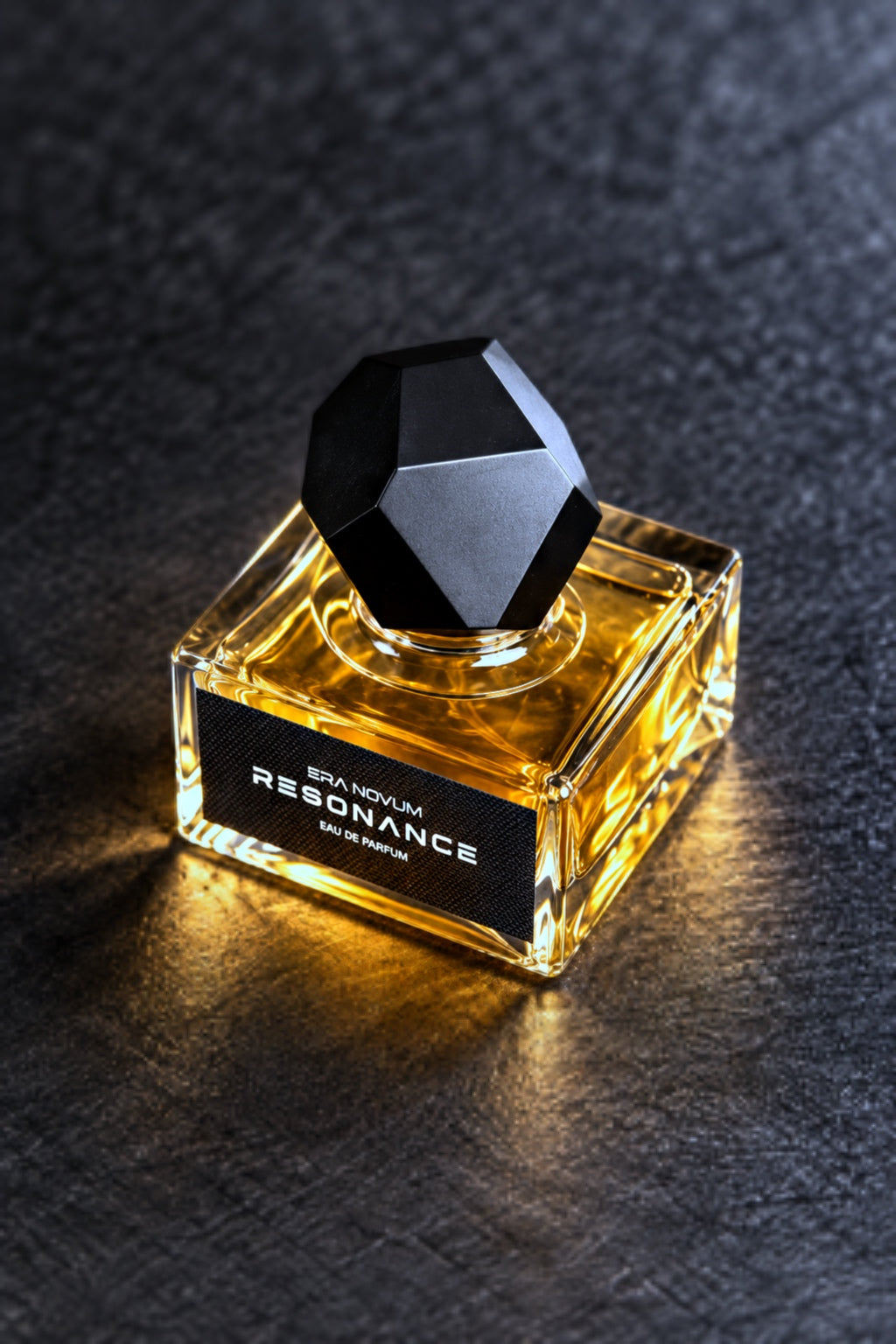 A bottle of RESONANCE Eau de Parfum with a faceted black cap and a black label, filled with amber-colored liquid, set on a dark textured surface.