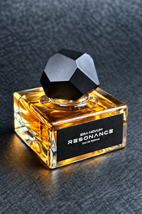 A bottle of Era Novum RESONANCE Eau de Parfum with amber-colored liquid in a clear square glass bottle, featuring a faceted black cap and a black label with white text, set against a dark textured background.